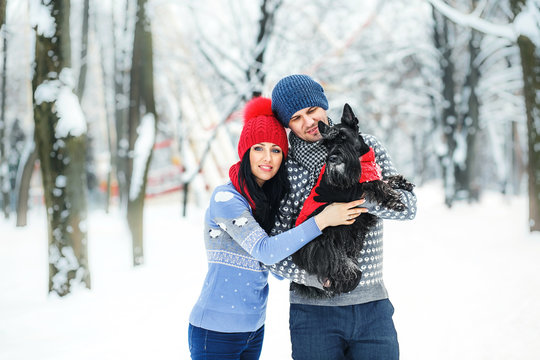 close up portrait of pretty young couple walking in winter park with dog scotch terrier in red hat and pullover. happy Valentine's Day together.