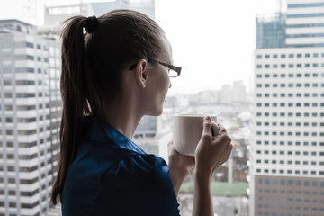 Businesswoman looking out her office window drinking coffee. 