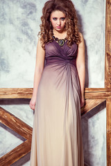 Obraz premium Fashion model with long dress and curly hairstyle and makeup posing near wall with wooden pole. studio shot. 
