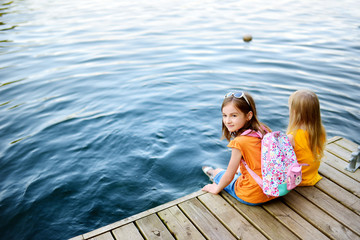 Two cute little girls sitting on a wooden platform by the river or lake