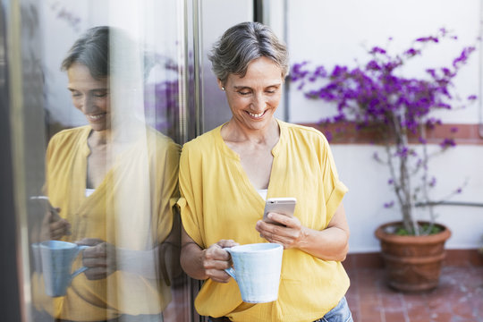 Happy mature woman using smart phone while holding coffee mug on porch