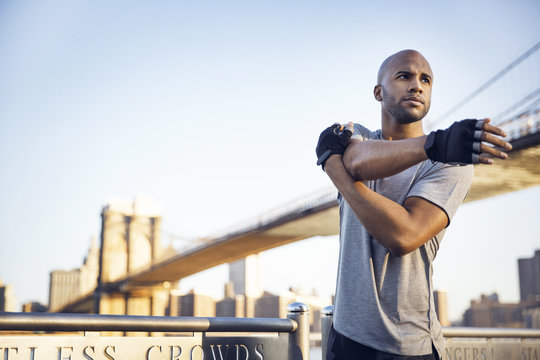 Determined male athlete doing stretching exercises on promenade with Brooklyn Bridge in background against clear sky