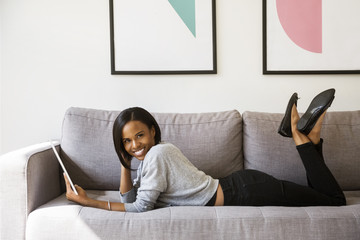 Side view portrait of happy woman holding tablet computer while lying on sofa at home