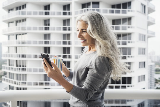 Happy mature woman using smart phone while holding coffee mug on balcony against building