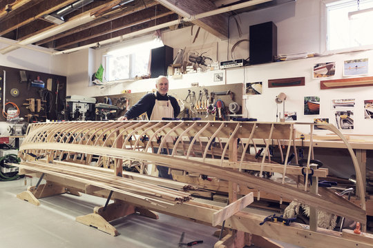 Portrait of confident senior man standing by incomplete boat in workshop