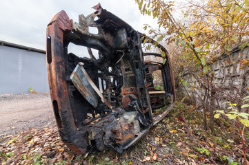 Rusty burned-out SUV on its side. Near the concrete fence on the background of trees and fallen leaves