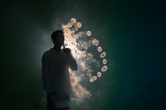 The Man Smoke An Electronic Cigarette With A Ring Against The Background Of The Dark Light