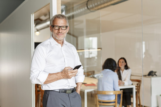 Confident businessman with cell phone in office with employees in background