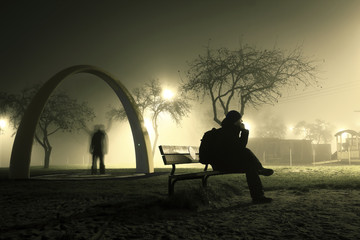 One man is sitting on the bench in foggy and mysterious park and ghost is behind him