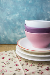 Stack of porcelane and ceramic plates and bowls on a wood kitchen table, floral linen cloth, blue and white concrete wall background, Provence rural interior