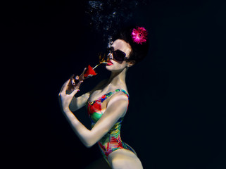 Obraz na płótnie Canvas beautiful girl in a colorful stylish swimsuit and sunglasses and pink gerbera in the head with a cocktail in her hand underwater in the swimming pool