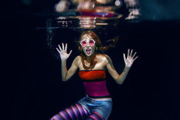 red hair funny girl in colorful clothes and pink sunglasses with tatto on the dark background swimming underwater with photo camera