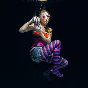 red hair funny girl in colorful clothes and pink sunglasses with tatto on the dark background swimming underwater with photo camera