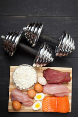 Proteins and dumbbells. Fish, cheese, eggs, meat, chicken  heart on a black wooden background - 134041029
