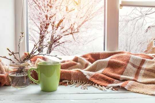 Cup of coffee, books, branch of cherry tree, wool blanket on windowsill. In the background snow tree pattern on window. Cozy home concept.
