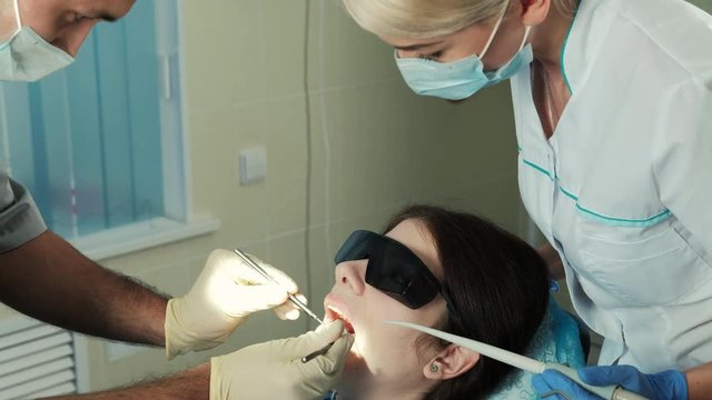 assistant oversees work treating of dentist in dental clinic office. young blonde woman working tooth doctor in mask looking for man by his actions carefully works treats teeth in protective masks