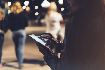 Girl pointing finger on screen smartphone on background illumination bokeh color light in night city, hipster using in hands and texting mobile phone, mockup glitter street, content lifestyle