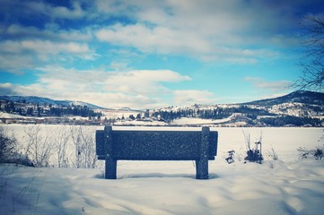 Scenic winter landscape view of frozen snow covered lake with closeup of park bench overlooking lake and mountains with bright blue sky and white clouds. 