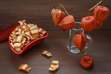 On a brown background physalis fruit in the vase and chips in the plate. The red triangular plate. Chips in the form of fried bacon.