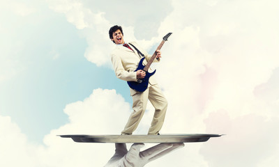 Businessman on metal tray playing electric guitar against blue sky background