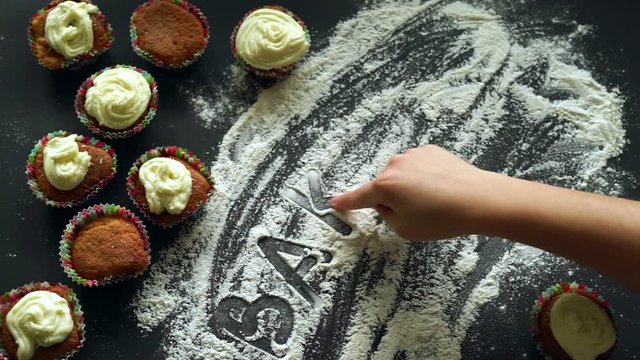 Baking concept. Finger writing word baking on flour at kitchen table. Inscription baking on white flour spills at table. Baked cupcakes on kitchen table