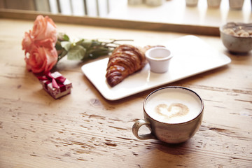 Obraz na płótnie Canvas Romantic breakfast, Valentine's Day celebrating. Present box, rose flowers, fresh croissant, coffee on wooden table. Focus on cup. Daylight from window.