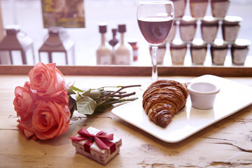 Romantic breakfast, Valentine's Day celebrating. Present box, rose flowers, fresh croissant, coffee on wooden table. Focus on cup. Daylight from window.