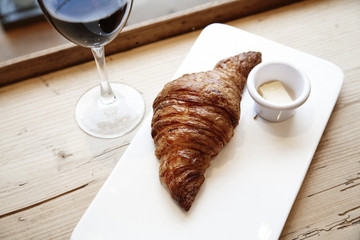Fresh bakery croissant and glass of red wine in wooden table. City cafe daylight near window