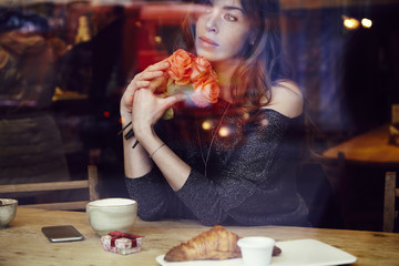 Beautiful woman with long hair holding red roses flowers in hand look through the window in cafe. Coffee and croissant on wooden table. St. Valentine's Day or Birthday concept.