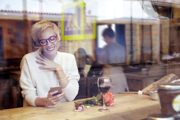 Beautiful blonde woman wearing eyeglasses messaging by mobile phone in cafe. Got a love message. Present box and rose flowers on wooden table. Romantic breakfast for a date or St. Valentine's Day. 