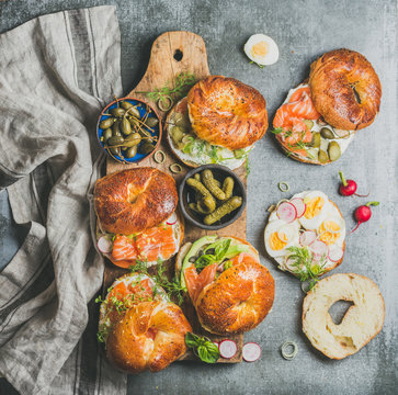 Variety of bagels with smoked salmon, eggs, radish, avocado, cucumber, greens and cream cheese for breakfast, healthy lunch, party or takeaway on wooden board over grey concrete background, top view