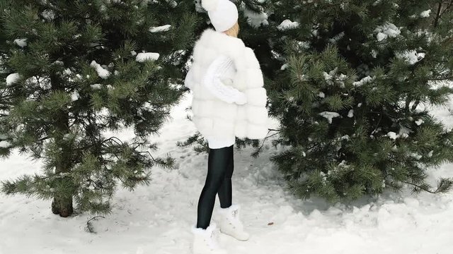 High model posing in a white dress in a green tree in the winter. The blonde woman wearing a white cap with pompons of different colors, a long woolen sweater, fur vest, gloves, boots with ornaments