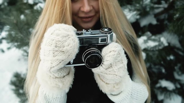 Tremendous girl shoots with film camera against background of winter landscape. Beautiful, charming blonde is dressed in white knitted sweater, black fur vest and pearl woolen mittens. She photographs