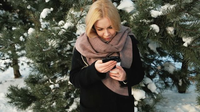 Beautiful woman checks messages near pines covered with snow. Fantastic blonde smiles for joy and happiness surprised and again laughs reading on screen of mobile phone amusing messages. She is