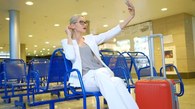 Passenger in waiting room at airport looking into phone indoors. Fashionable blond woman in white suit, grey blouse, black spectacles. Nice female makes selfie on telephone before travelling, waiting