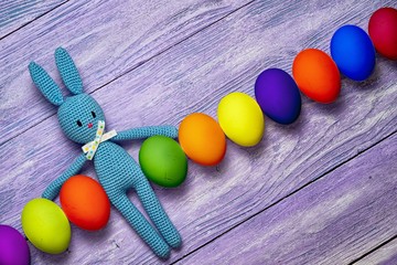 Amigurumi toy easter bunny with diagonal row of colorful eggs on a wooden table - easter background with copy space