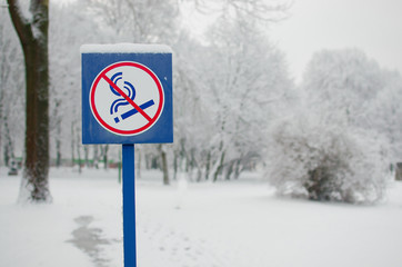 No smoking metal sign pole in the winter park. Don't smoke sign. Blue no smoking label stick to the pole in the located public park with winter background. Public park in the city for health lovers.