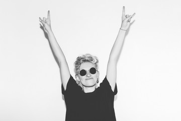 Stylish fashion sexy blonde bad girl in a black t-shirt and rock sunglasses. Dangerous rocky emotional woman giving the   Roll sign, devil horns gesture.   white toned.