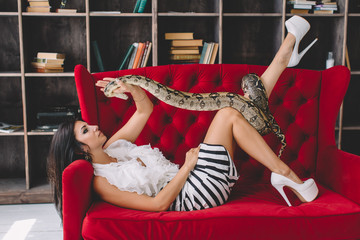 Young brunette posing in bright studio and laying on red sofa with a snake python 
