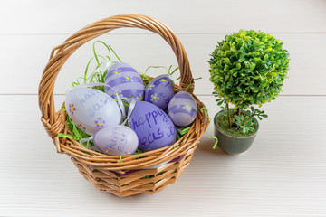 Easter wicker basket with colored eggs and a small bonsai on white wooden board.
