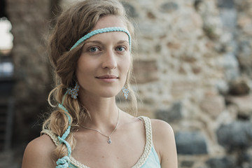 Close up portrait of beautiful blonde girl with blue eyes and braid wearing blue earrings and turquoise headband against blurred background of stone wall