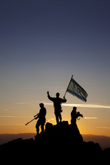 Three armed soldiers with the Israeli flag on the top of the mountain