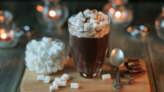 Beautiful composition - hot chocolate with marmalade and chocolate pieces in a transparent glass. The glass stands on a wooden stand. Behind the candles are burning