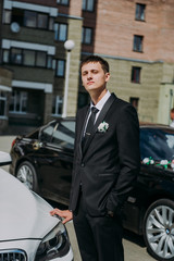 stylish elegant groom holding bouquet standing near car on the background of old city