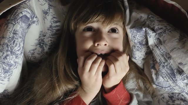 A little girl lying in her bed, under the sheets, at night. She is alarmed and scared.
