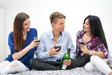 Three young friends sitting on bed and drinking red wine together.