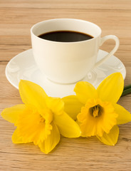 A cup of coffee with two daffodils in the foreground on a brown wooden background.
