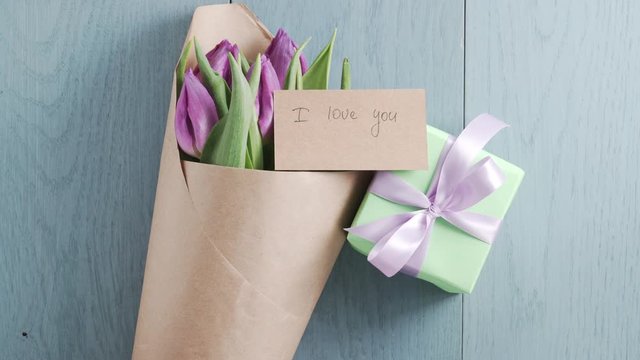 man puts i love you greeting card on purple tulips bouquet with gift on blue table shot from above, uhd prores footage