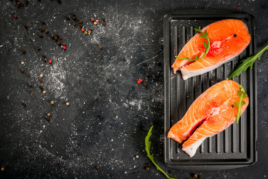 Raw uncooked salmon steaks on the boards for the grill, on a dark gray stone kitchen table. With salt, spices, seasonings, herbs and vegetables for cooking.  Top view, copy space