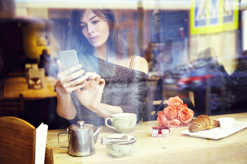 Beautiful caucasian woman with long hair texting message by mobile phone, sitting in cafe. Focus on hands. Romantic breakfast for a date or St. Valentine's Day. Present box and rose flowers. 